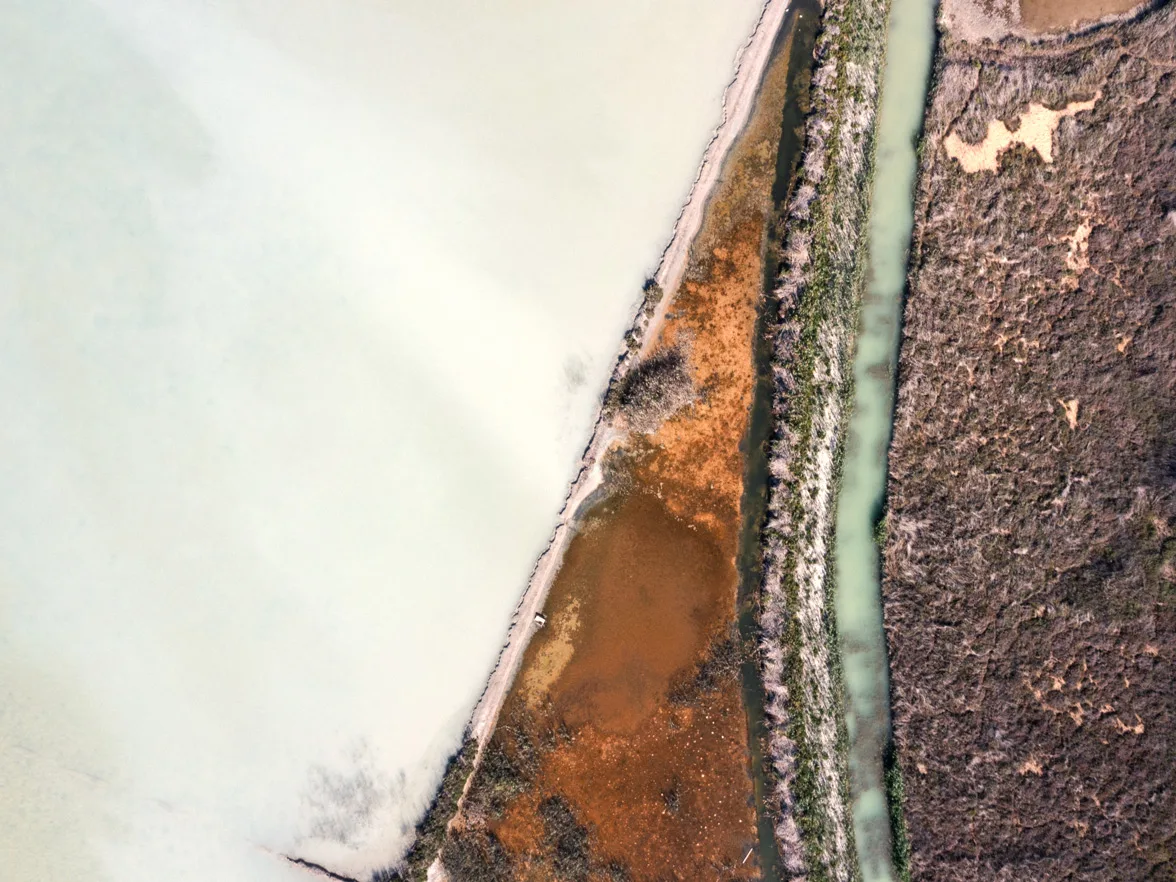 Aerial view shows a turquoise canal separating light beige land from reddish-brown, dry vegetation under bright daylight. Aerial view of a turquoise canal winding through brown earth and vegetation near Calpe Sapin, limited edition fine art print by Ludwig Favre