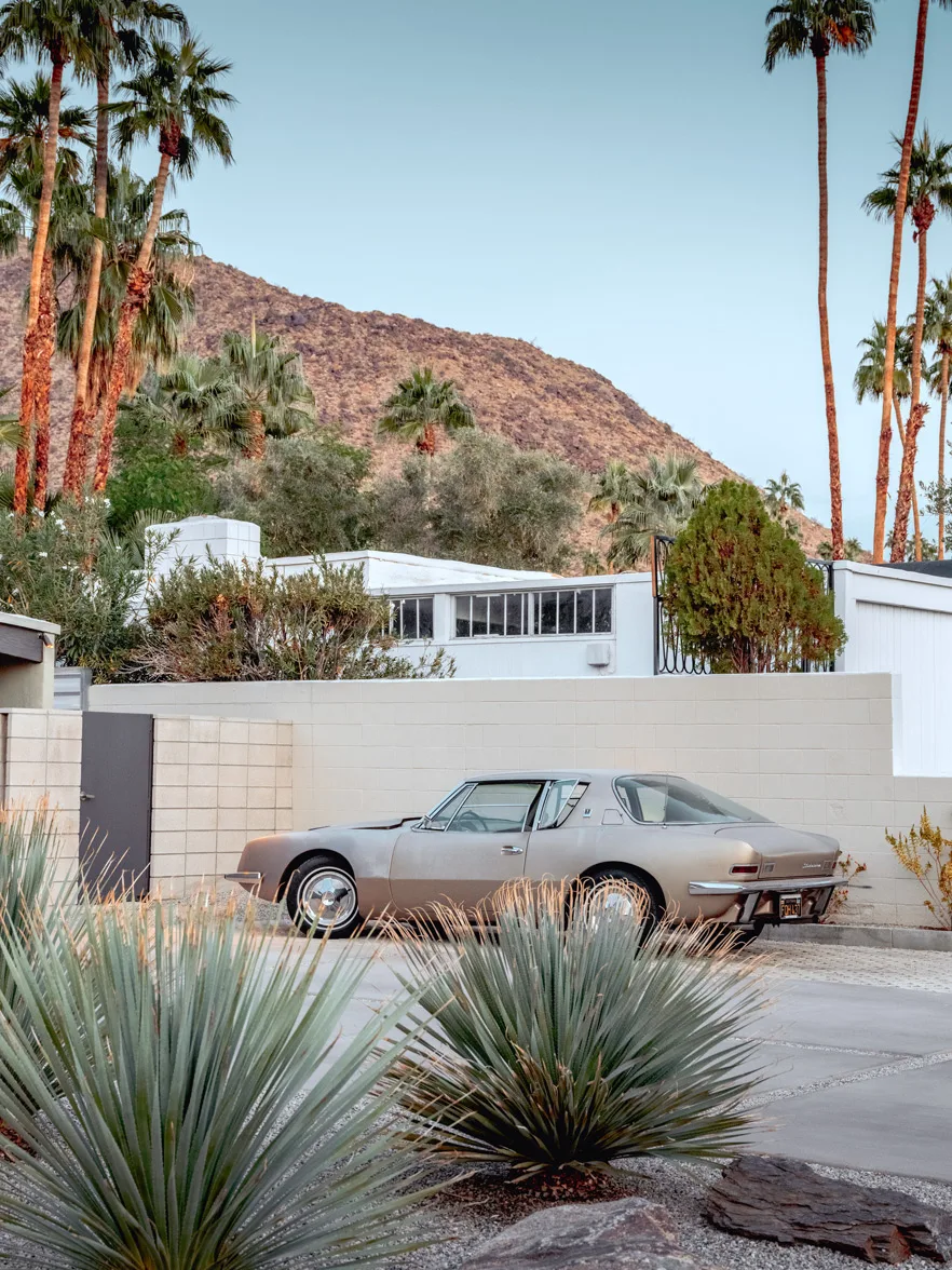 A vintage silver car sits on a driveway in Palm Springs, flanked by agave plants and palm trees against a backdrop of mountains at dusk. Vintage car parked on a midcentury driveway in Palm Springs, California, with desert landscaping and the San Jacinto mountains, limited edition fine art print by Ludwig Favre