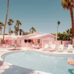 Pink motel poolside with lounge chairs, palm trees and colorful floaties under clear blue sky.