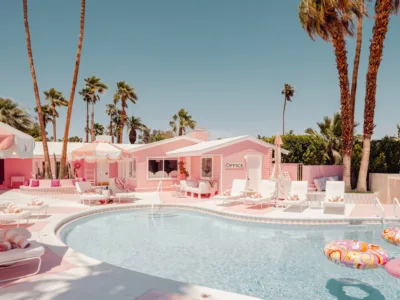 Pink motel poolside with lounge chairs, palm trees and colorful floaties under clear blue sky.