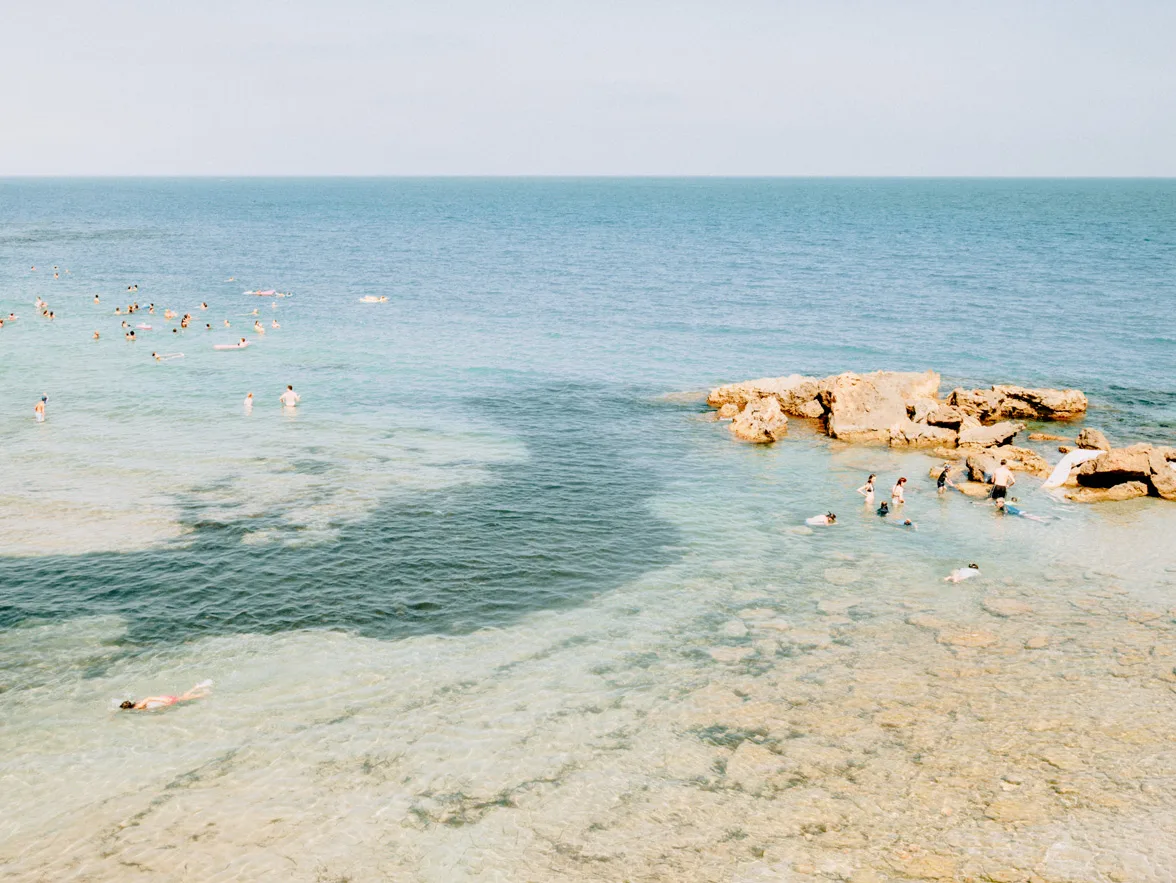 Swimmers in clear blue water with varying shades near rocky outcrops on a sandy beach under sunny summer skies. Swimmers around rocky outcrops in clear Mediterranean waters under soft summer light, limited edition fine art print by Ludwig Favre