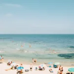 Sunbathers and swimmers on a sandy beach with colorful umbrellas under clear blue sky.