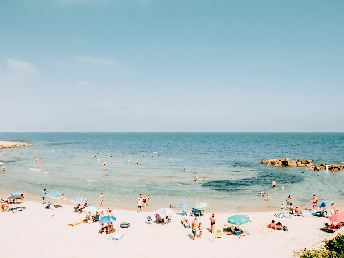 People swimming, sunbathing, and walking on sandy beach under striped umbrellas at Costa del Sol, clear blue sky, bright sunlight with rocky outcrops in the sea Striped umbrellas on a sandy beach at Costa del Sol under clear blue sky, limited edition fine art print by Ludwig Favre