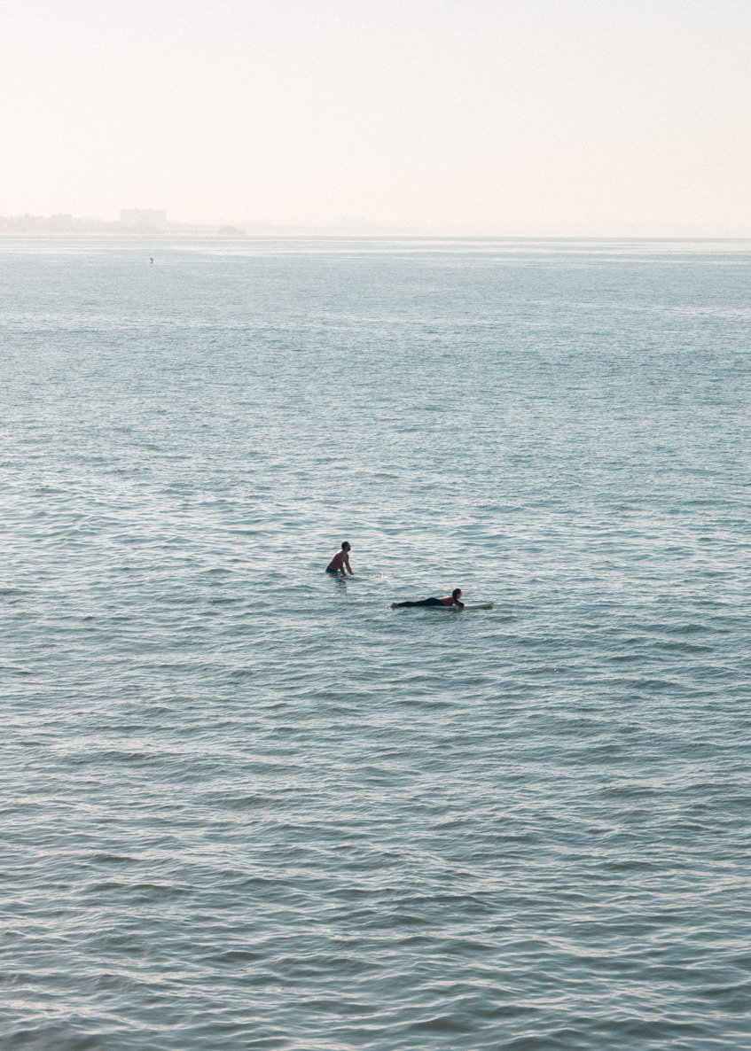 Surfer riding a small wave, a person floating on their board in calm blue-gray ocean under overcast sky. A surfer riding a wave in the Pacific Ocean off Los Angeles at soft morning light, limited edition fine art print by Ludwig Favre