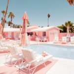 Pink umbrellas surround a pool in a bright, pink resort with palm trees under a clear blue sky.