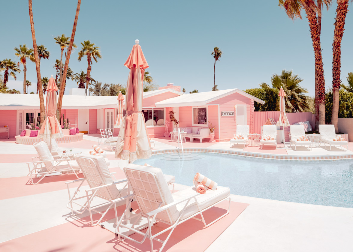 Pink umbrellas surround a pool in a bright, pink resort with palm trees under a clear blue sky.