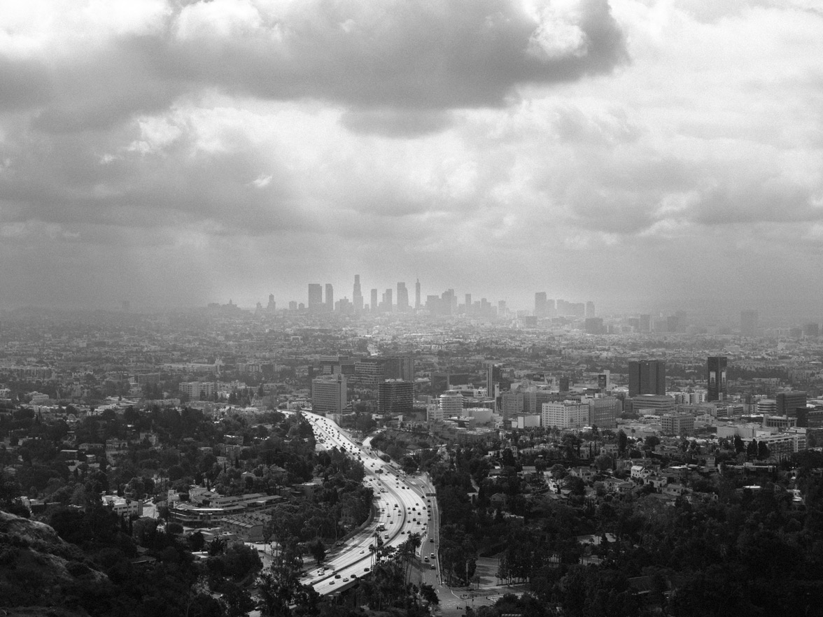 Black and white view of downtown Los Angeles with a winding highway cutting through the city, seen from the Hollywood Hills under an overcast sky.