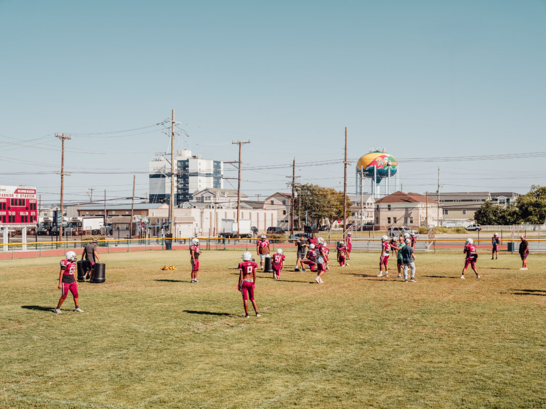 Athletes in red football gear stand on a sunny grass field with an urban skyline and colorful water tower behind them.