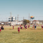Athletes in red football gear stand on a sunny grass field with an urban skyline and colorful water tower behind them.