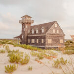 A weathered wooden station with a watchtower stands among sandy dunes under a soft, golden-hour sky.