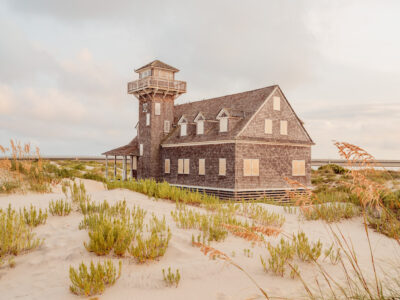 A weathered wooden station with a watchtower stands among sandy dunes under a soft, golden-hour sky.