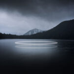 A bright circular neon light reflects on a dark, still mountain lake surrounded by dense trees under a cloudy night sky with mountains in the distance.