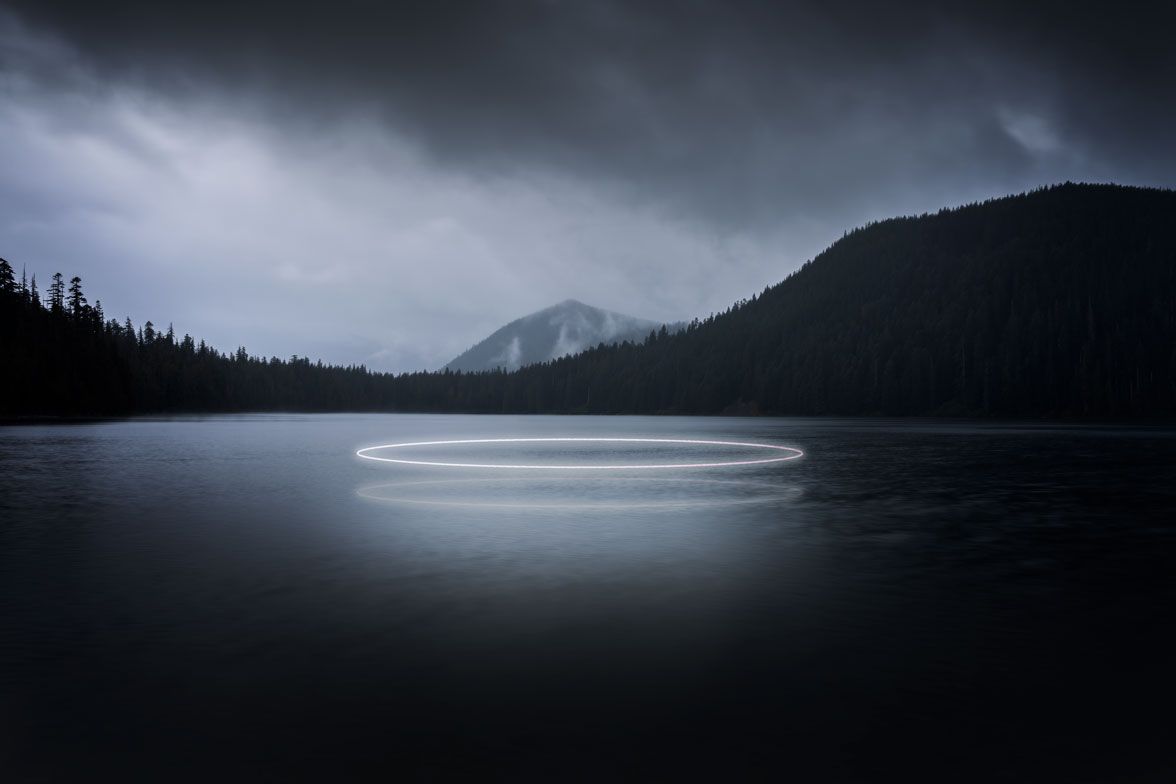 A bright circular neon light reflects on a dark, still mountain lake surrounded by dense trees under a cloudy night sky with mountains in the distance.