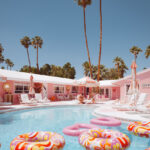 Pink motel courtyard with colorful pool floats, palm trees and lounge chairs under bright blue sky.