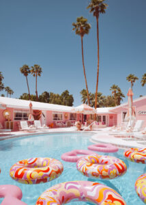 Pink motel courtyard with colorful pool floats, palm trees and lounge chairs under bright blue sky.