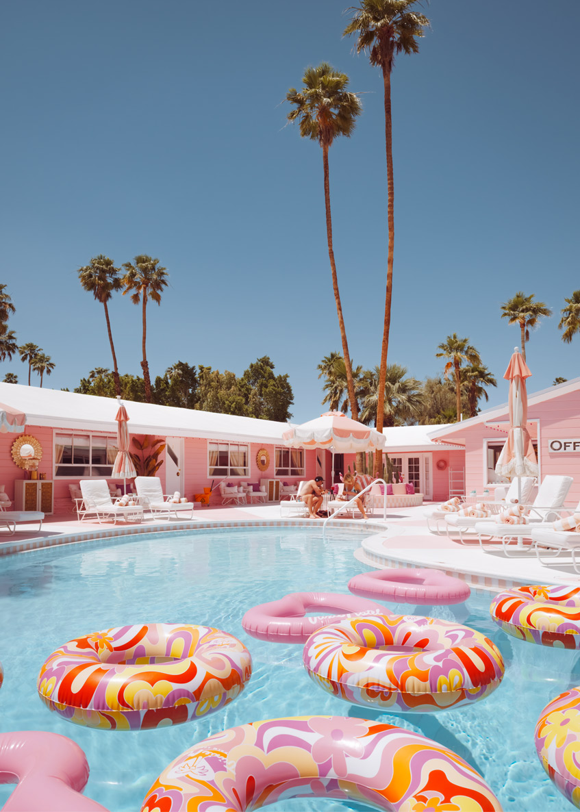 Pink motel courtyard with colorful pool floats, palm trees and lounge chairs under bright blue sky.