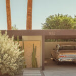 Vintage silver car parked under a tan mid-century carport with palm trees and cacti in bright sunlight.