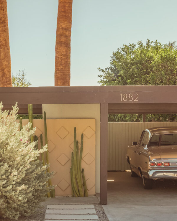 Vintage silver car parked under a tan mid-century carport with palm trees and cacti in bright sunlight.