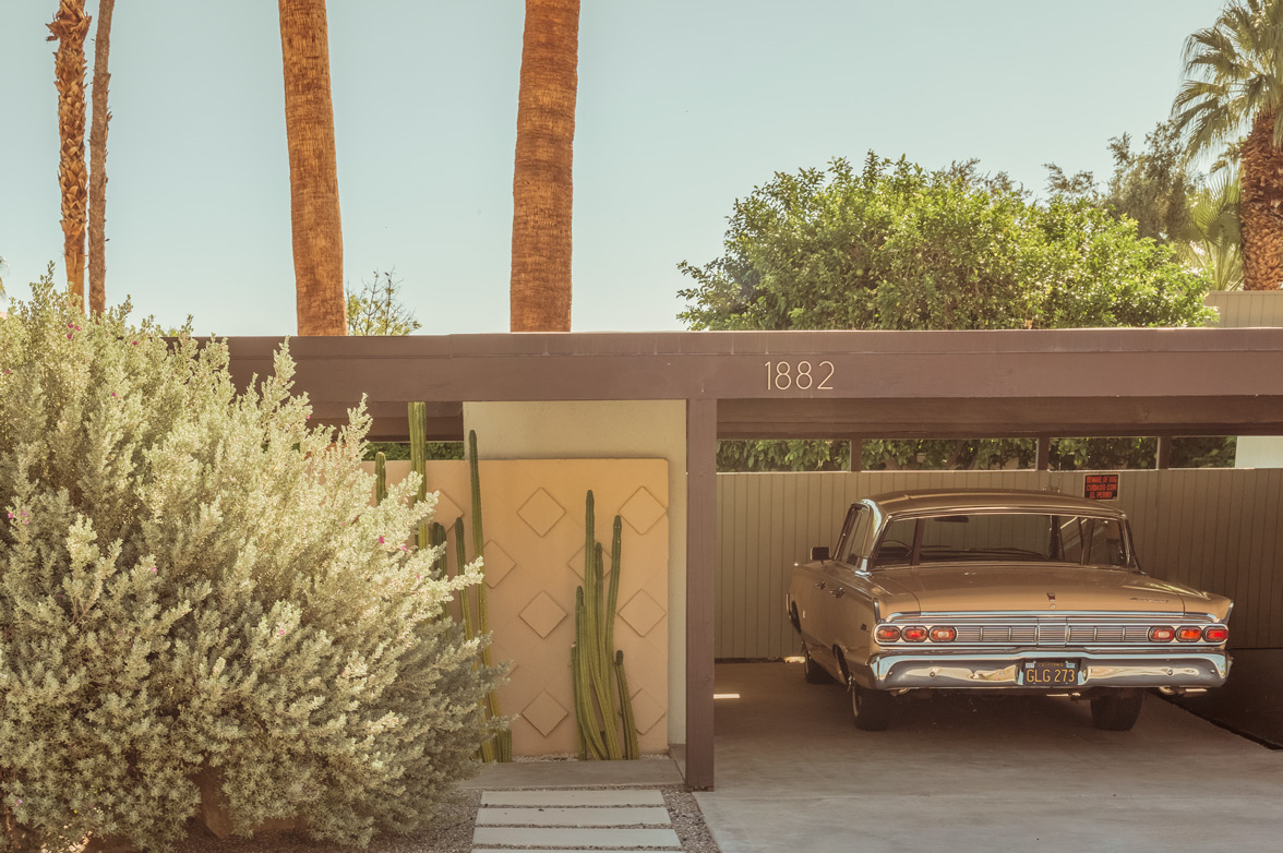 Vintage silver car parked under a tan mid-century carport with palm trees and cacti in bright sunlight.