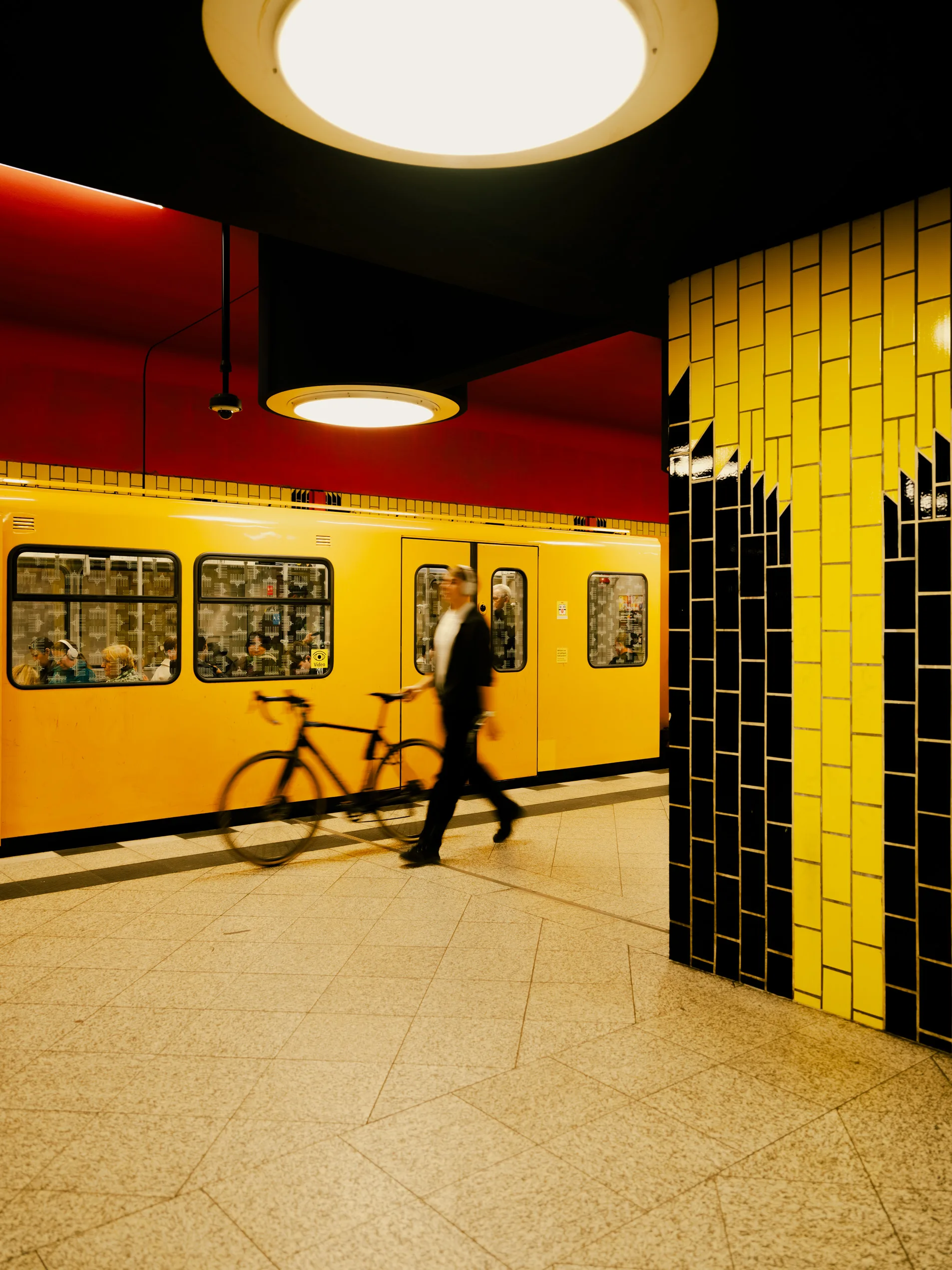 A blurred man in dark clothes pulls a bicycle past a yellow subway train in a red-and-yellow tiled Berlin station. A blurred man in dark clothes pulls a bicycle past a yellow subway train in a red-and-yellow tiled Berlin station.