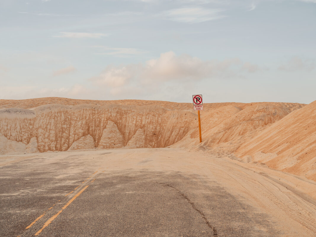 Red no parking sign amidst pale orange sand dunes and a desert road, fine art print by Ludwig Favre