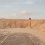 Red no parking sign amidst pale orange sand dunes and a desert road, fine art print by Ludwig Favre