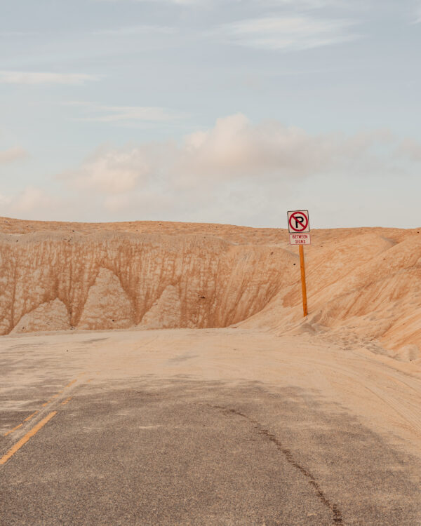 Red no parking sign amidst pale orange sand dunes and a desert road, fine art print by Ludwig Favre