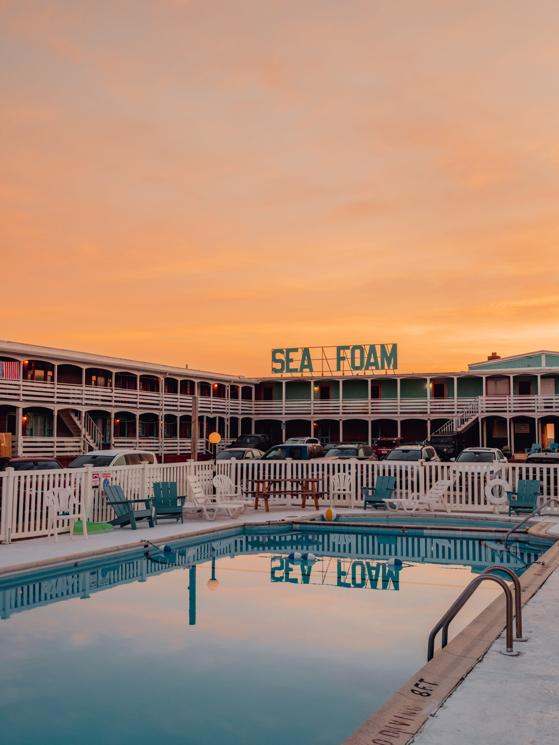 A pool reflects a warm orange sunset sky in front of the two-story Sea Foam Motel and its turquoise sign. Turquoise swimming pool reflecting teal Sea Foam Motel sign under an orange sky in the Outer Banks, fine art print by Ludwig Favre