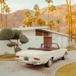 White vintage Mercedes parked in a driveway before a modern house, palm trees, and mountains in warm sunset light.
