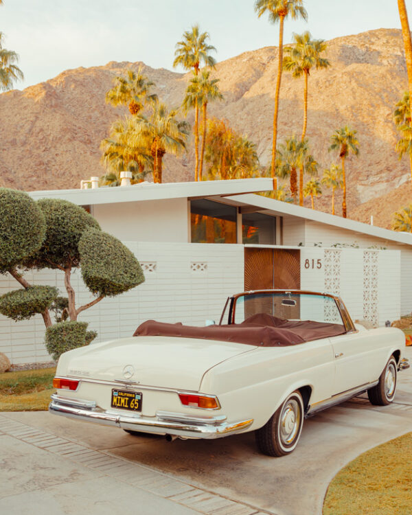 White vintage Mercedes parked in a driveway before a modern house, palm trees, and mountains in warm sunset light.