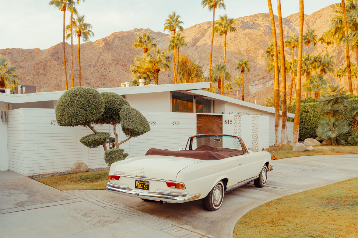 White vintage Mercedes parked in a driveway before a modern house, palm trees, and mountains in warm sunset light.