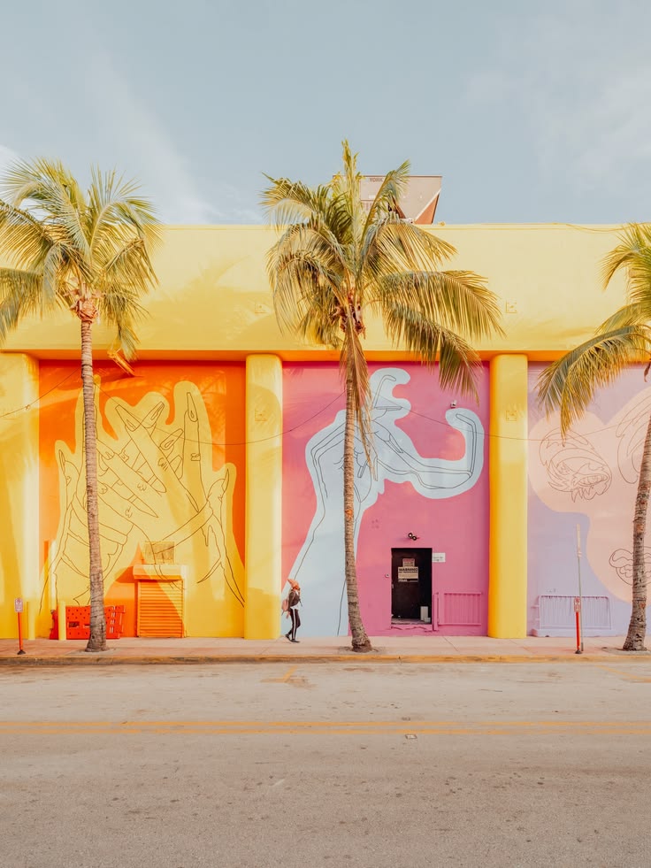 A person walks past a Miami wall of pink, orange, and lavender panels with hand murals, framed by three palm trees. Miami pink wall print: pink, orange and lavender panels with hand murals and three palm trees