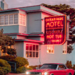 A vintage red car parked in front of a pink and blue motel with a neon "Ocean Park Motel" sign under a soft pastel sky.