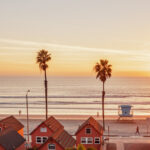 Sunset over Oceanside beach with palm trees, a lifeguard tower, and small houses in warm orange and pink tones.