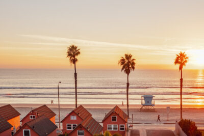 Sunset over Oceanside beach with palm trees, a lifeguard tower, and small houses in warm orange and pink tones.